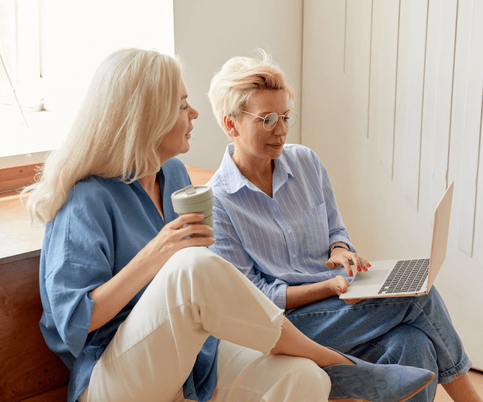 Two women sitting on a sofa with a laptop and a mug